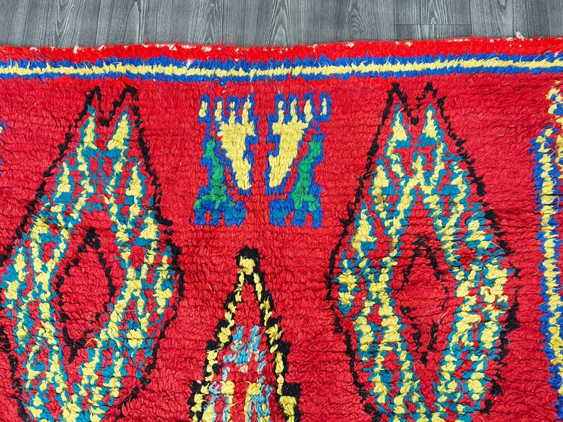 May include: A red, hand-woven rug with a geometric pattern in yellow, blue, and green. The rug has a fringe border.