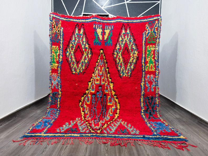 May include: A red Moroccan rug with a geometric pattern in shades of blue, yellow, and green. The rug has a fringe border and is woven with a thick, textured pile.