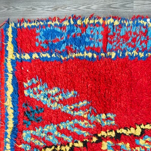 May include: A red, blue, and yellow hand-woven rug with a fringe border. The rug has a geometric pattern with a central design of stylized flowers.