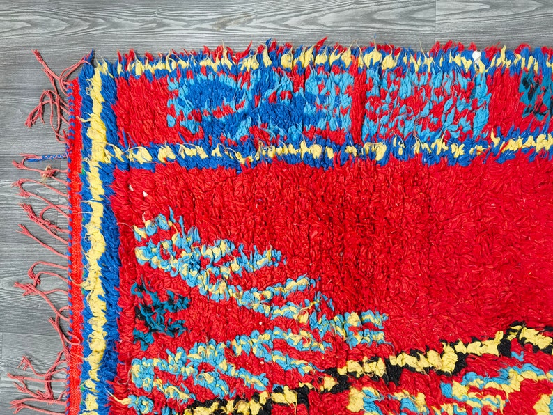 May include: A red, blue, and yellow hand-woven rug with a fringe border. The rug has a geometric pattern with a central design of stylized flowers.