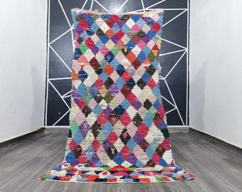 May include: A colorful Moroccan rug with a diamond pattern in shades of pink, blue, red, and purple. The rug has a white background and is made of wool.
