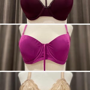 Op de afbeelding: Drie bh's tentoongesteld op mannequins. De bovenste bh is bordeauxrood met een delicate kantversiering en een kruisbandontwerp. De middelste bh is fel fuchsia met een eenvoudig ontwerp. De onderste bh is lichtbeige met een kantversiering en een satijnen afwerking.