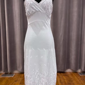 May include: A white slip dress with adjustable straps and floral embroidery. The bodice has a V-neck design with delicate floral lace detailing. The skirt features embroidered floral accents near the hem, creating a vintage look.