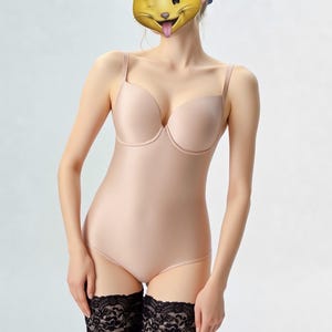 Puede incluir: Un body color nude con tirantes finos y sujetador incorporado. La modelo lleva medias negras con encaje en la parte superior. El body es de color liso y parece estar hecho de un material suave y ajustado.