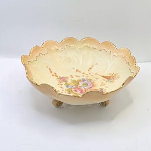 May include: A decorative, scalloped-edge bowl with a floral design. The bowl has a cream-colored interior and a peach-colored exterior with gold trim. The floral design includes pink, blue, and yellow flowers. The bowl is supported by three small feet.