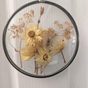 May include: A round, clear glass ornament with a black metal frame, filled with dried yellow flowers and stems. A purple ribbon is attached for hanging. The flowers have a natural, rustic appearance, creating a decorative piece.