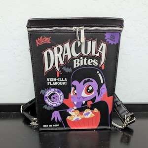 May include: A black structured backpack shaped like a cereal box with a silver zipper closure. The front features a graphic of a vampire with the text "Dracula Bites," "Vein-illa Flavour!," "Kill Your Hunger," and "Net Wt 666g" printed on it.