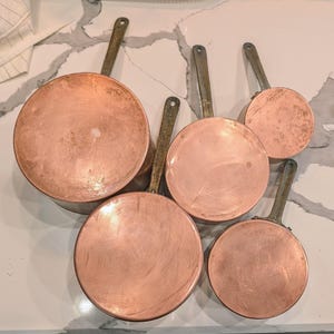 May include: A collection of five copper pans with brass handles. The pans vary in size, with a large pan at the top left and smaller pans arranged below and to the right. The copper surfaces have a warm, reddish-brown hue, and the handles are a muted gold color.