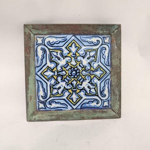 May include: A square ceramic tile featuring a symmetrical floral and geometric pattern in shades of blue, white, and yellow. The tile is encased in a weathered, oxidized metal frame with a textured, greenish-brown patina finish.