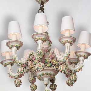 May include: A multi-armed chandelier featuring white ceramic construction with hand-painted pink rose motifs, green leaves, and yellow floral accents. The fixture includes six white fabric lamp shades and a central hanging ceramic finial.