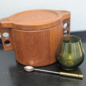 May include: A wooden ice bucket with a lid, a green glass, and a silver spoon with a gold handle. The ice bucket is made of vertical wooden planks and has a handle with two circular cutouts. The green glass has a rounded shape.