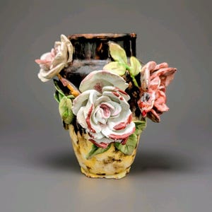 May include: A ceramic vase with a dark brown top and a yellow base, adorned with sculpted roses in shades of white, pink, and red, along with green leaves. The vase has a textured appearance, suggesting a handcrafted design.
