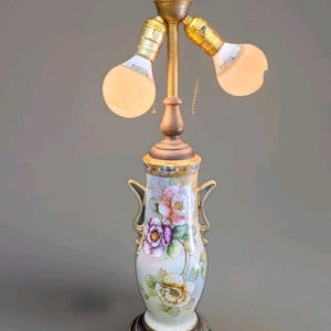Peut inclure: Une lampe de table avec une base en porcelaine en forme de vase ornée de motifs floraux peints à la main en rose, violet et jaune. La lampe possède deux douilles en laiton avec des ampoules allumées, des anses dorées et une base en métal texturé.