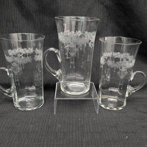 May include: A set of three clear glass mugs with handles, each featuring a delicate etched floral pattern around the upper rim. The central mug is elevated on a small, clear acrylic stand, while the two flanking mugs sit at a lower level.