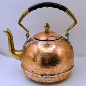May include: A copper tea kettle with a hammered texture, brass accents, and a black handle. The spout and handle supports are brass. The lid is topped with a decorative finial. The kettle is designed for stovetop use.