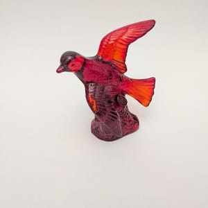 Peut inclure: Une figurine en verre rouge translucide représentant un oiseau en plein vol, posé sur une base texturée. L'oiseau présente des tons cramoisi profonds, un bec pointu et des ailes déployées avec des détails de plumes moulés visibles.