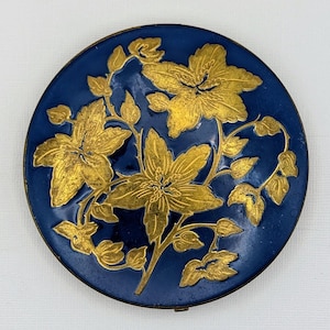 Op de afbeelding: Een ronde, marineblauwe compact met een gouden bloemenontwerp. De gouden bloemen en bladeren zijn gedetailleerd tegen de donkerblauwe achtergrond. De compact heeft een metalen rand en een scharnierende opening.