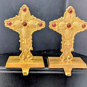 Puede incluir: Dos colgadores de calcetines de metal dorado ornamentados en forma de cruces. Cada cruz presenta detalles de joyas rojas y descansa sobre una base rectangular con un gancho para colgar calcetines. Las cruces tienen diseños decorativos detallados.