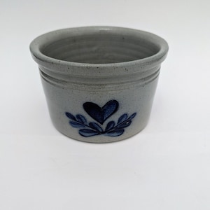 Puede incluir: Pequeño cuenco redondo de gres con un esmalte azul grisáceo claro. Un diseño de corazón y hojas azul oscuro está centrado en la parte delantera. El cuenco tiene una superficie ligeramente texturizada y un diseño sencillo y clásico.