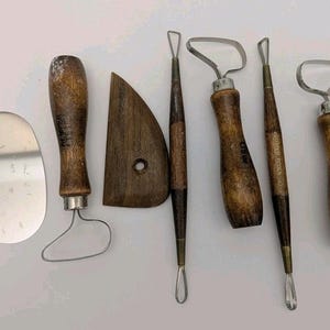 May include: A collection of pottery tools with wooden handles and metal blades. The set includes a metal rib, a wooden kidney tool, and various loop tools. The handles are a dark brown color, and the metal parts are silver.