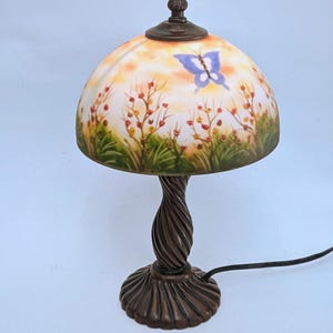 Può includere: Lampada da tavolo decorativa con paralume in vetro dipinto con un design a farfalla e floreale. Il paralume è a forma di cupola, appoggiato su uno stelo e una base tortuosi color bronzo. La lampada ha un terminale marrone scuro e un cavo di alimentazione nero.