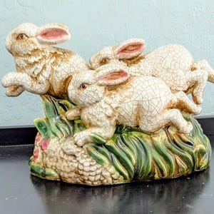 May include: A ceramic figurine featuring three rabbits in mid-leap, atop a base of green grass and pink flowers. The rabbits have a cracked, aged appearance with pink-lined ears and red eyes. The figurine is a decorative item.