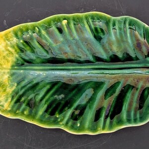 May include: A ceramic leaf-shaped dish with a glossy finish. The dish features a gradient of colours, transitioning from yellow to green, with brown accents. The leaf has a handle on one end and is designed with vein-like details.