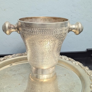 May include: A silver ice bucket with two ornate handles, resting on a matching silver tray. The bucket has a textured surface with a floral pattern. The tray has a decorative, scalloped edge.