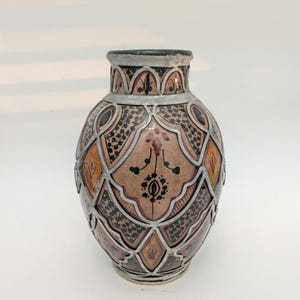 May include: A ceramic vase featuring a tan and brown base with intricate black painted patterns and geometric shapes. The exterior is overlaid with a silver-toned metal filigree framework that outlines the decorative panels and the rim.