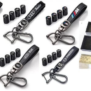 May include: A selection of black keyrings featuring car brand emblems such as Mercedes-Benz, Audi, BMW, Hyundai, Kia, and Mazda. Each keyring has a black leather strap and a metal clasp. Also shown are black tyre valve stem caps.