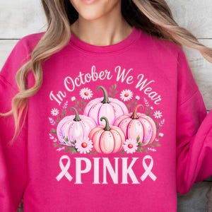 Könnte beinhalten: Ein magentafarbenes Sweatshirt mit einer Grafik aus rosa Kürbissen, Blumen und dem Text "In October We Wear PINK" mit einem rosa Band. Das Design ist auf der Vorderseite des langärmeligen Rundhalsausschnitts zentriert.