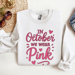 Könnte beinhalten: Weißes Sweatshirt mit dem pinkfarbenen Text "IN October we wear Pink". Das Design umfasst rosa Herzen und Punkte. Das Sweatshirt wird auf einer weißen Oberfläche mit Sonnenbrille und einem geflochtenen Korb im Hintergrund präsentiert.