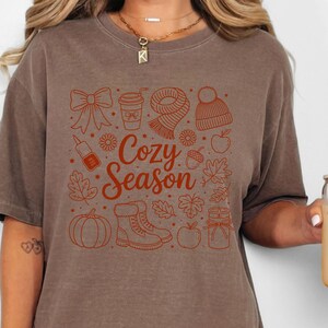 K&ouml;nnte beinhalten: Braunes T-Shirt mit einem herbstlichen Motiv. Das Design umfasst eine Schleife, eine Kaffeetasse, einen Schal, eine M&uuml;tze, Bl&auml;tter, einen K&uuml;rbis, Stiefel, einen Apfel und die Worte "Cozy Season".