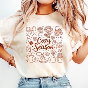 K&ouml;nnte beinhalten: Cremefarbenes T-Shirt mit brauner Grafik mit herbstlichen Illustrationen. Das Design umfasst eine Schleife, eine Kaffeetasse, einen Schal, eine M&uuml;tze, einen K&uuml;rbis, Stiefel, Bl&auml;tter und den Text "Cozy Season."