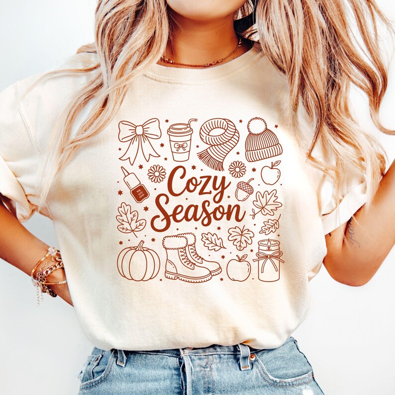 K&ouml;nnte beinhalten: Cremefarbenes T-Shirt mit brauner Grafik mit herbstlichen Illustrationen. Das Design umfasst eine Schleife, eine Kaffeetasse, einen Schal, eine M&uuml;tze, einen K&uuml;rbis, Stiefel, Bl&auml;tter und den Text "Cozy Season."