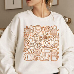 Könnte beinhalten: Weißes Sweatshirt mit braunem Herbst-Design. Die Grafik zeigt einen Kürbis, Blätter und einen Stiefel, mit dem Text "Tis the Season" in einer dekorativen Schriftart.