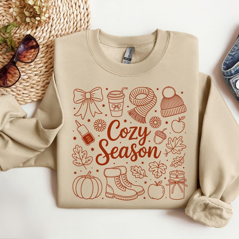 K&ouml;nnte beinhalten: Beiges Sweatshirt mit herbstlichem Design. Das Design umfasst eine Schleife, Kaffeetasse, Schal, M&uuml;tze, Nagellack, Blumen, Bl&auml;tter, K&uuml;rbis, Stiefel, &Auml;pfel und die Worte "Cozy Season" in Rotbraun.