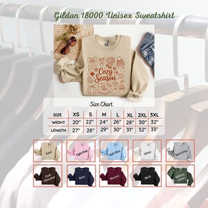 K&ouml;nnte beinhalten: Beiges Gildan 18000 Unisex-Sweatshirt mit dem Schriftzug "Cozy Season" und herbstlichen Grafiken. Das Bild enth&auml;lt eine Gr&ouml;&szlig;entabelle und Farboptionen wie Sand, Rosa, Blau und Schwarz.