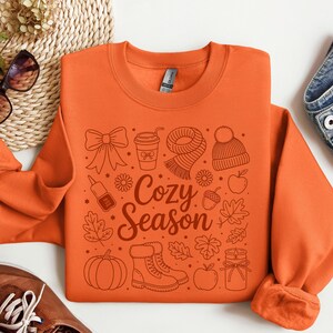 K&ouml;nnte beinhalten: Orangenes Sweatshirt mit einem grafischen Design mit herbstlichen Illustrationen, darunter eine Schleife, eine Kaffeetasse, ein Schal, eine M&uuml;tze, Bl&auml;tter, ein K&uuml;rbis, Stiefel und &Auml;pfel. Der Text "Cozy Season" befindet sich in der Mitte.