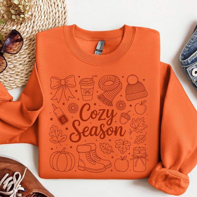 K&ouml;nnte beinhalten: Orangenes Sweatshirt mit einem grafischen Design mit herbstlichen Illustrationen, darunter eine Schleife, eine Kaffeetasse, ein Schal, eine M&uuml;tze, Bl&auml;tter, ein K&uuml;rbis, Stiefel und &Auml;pfel. Der Text "Cozy Season" befindet sich in der Mitte.