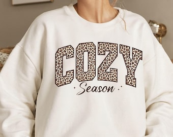 May include: A white sweatshirt with the words "COZY Season" printed on the front. The text "COZY" is in a brown leopard print font, and "Season" is in a cursive font. The sweatshirt is a crew neck style.