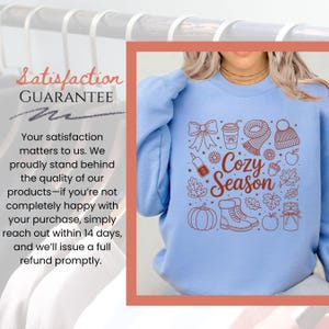K&ouml;nnte beinhalten: Hellblaues Sweatshirt mit einem braunen Grafikdesign mit herbstlichen Elementen wie einem K&uuml;rbis, Bl&auml;ttern und einem Schal. Der Text "Cozy Season" ist ebenfalls enthalten. Das Bild enth&auml;lt auch den Text "Satisfaction Guarantee".
