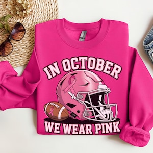Könnte beinhalten: Ein leuchtend pinkfarbenes Sweatshirt mit dem Text "IN OCTOBER WE WEAR PINK". Das Design umfasst einen pinkfarbenen Footballhelm und einen Football, beide mit rosa Schleifenakzenten. Das Sweatshirt hat einen Rundhalsausschnitt.