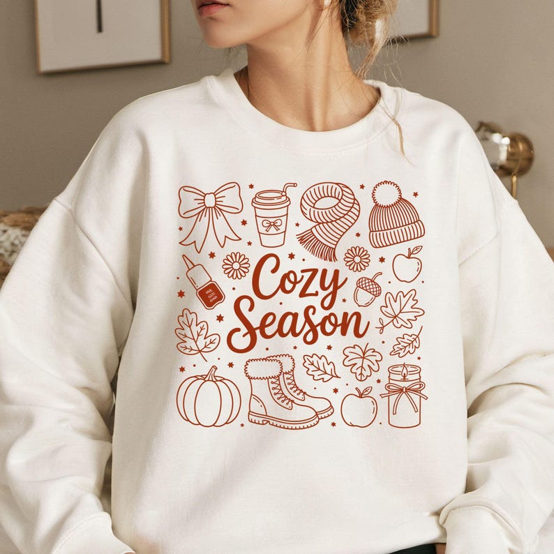 K&ouml;nnte beinhalten: Wei&szlig;es Sweatshirt mit braunem Grafikdesign. Das Design umfasst eine Schleife, eine Kaffeetasse, einen Schal, eine M&uuml;tze, einen Apfel, Bl&auml;tter, einen K&uuml;rbis, Stiefel und den Text "Cozy Season".
