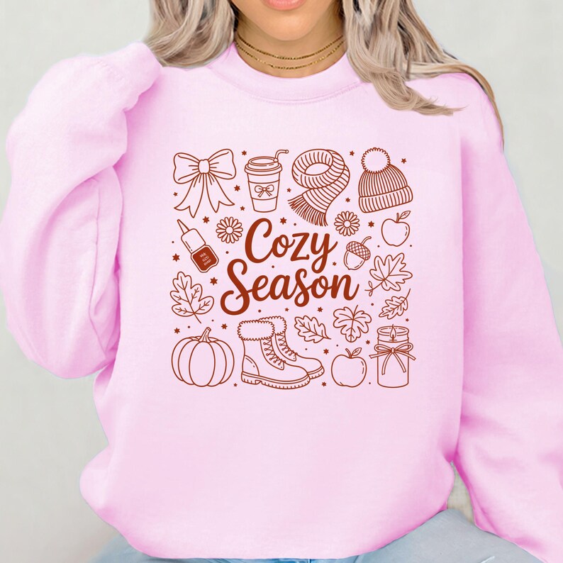 K&ouml;nnte beinhalten: Ein hellrosa Sweatshirt mit einem braunen Grafikdesign mit herbstlichen Illustrationen. Das Design umfasst eine Schleife, eine Kaffeetasse, einen Schal, eine M&uuml;tze, Nagellack, Blumen, Bl&auml;tter, einen K&uuml;rbis, Stiefel, &Auml;pfel, Eicheln und eine Kerze. Die Worte "Cozy Season" stehen im Zentrum.