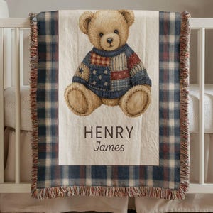 Puede incluir: Una manta para bebé beige con una ilustración de oso de peluche con un suéter de retazos. La manta tiene un borde a cuadros azul, rojo y blanco y el nombre "HENRY James" impreso en negro.