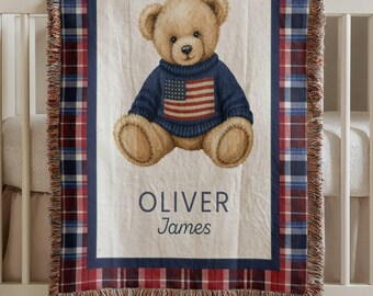 May include: A woven throw blanket featuring a central illustration of a light brown teddy bear wearing a navy blue sweater with an American flag design. The name OLIVER James is printed below the bear. The blanket has a red, white, and blue plaid border.