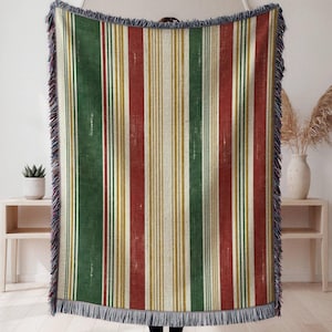 May include: A woven throw blanket featuring a vertical striped pattern in shades of forest green, deep red, and mustard yellow, accented by thin white lines. The rectangular textile is finished with a decorative fringe along the top and bottom edges.