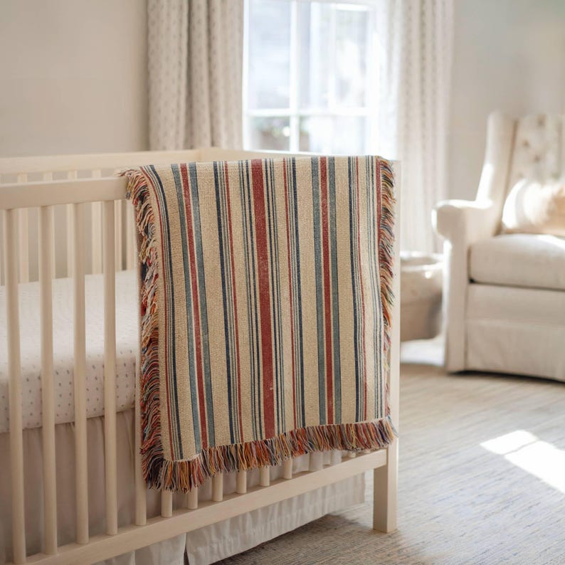 May include: A woven throw blanket featuring a vertical striped pattern in shades of cream, red, and blue, finished with decorative fringe along the top and bottom edges, draped over the side of a light-colored wooden crib with slatted sides.