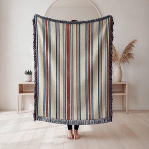 May include: A woven throw blanket featuring a vertical striped pattern in shades of cream, deep red, and muted blue. The blanket has a textured appearance with frayed fringe edges along the top and bottom, creating a classic textile design.